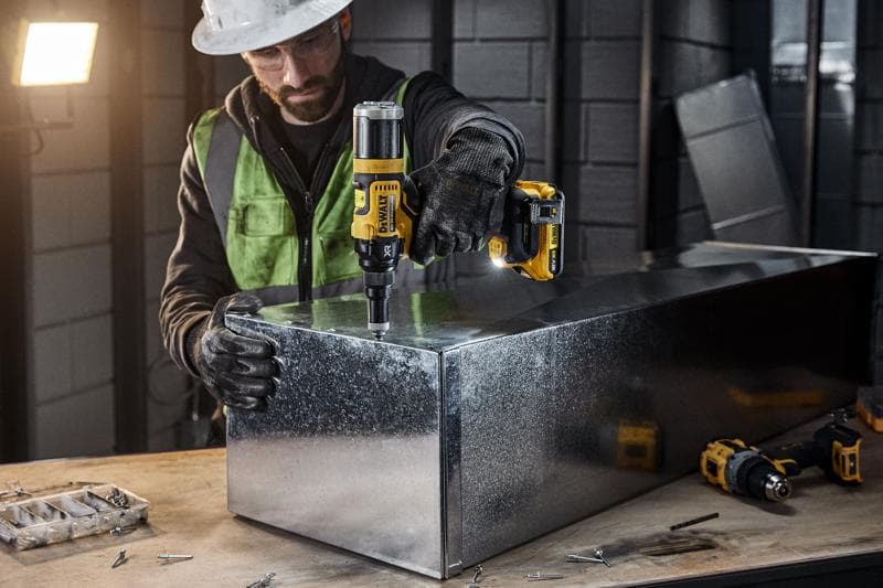 Une personne équipée d'un équipement de sécurité utilise un outil électrique sans fil DEWALT DCF403NT pour travailler sur une boîte en métal dans un atelier industriel. Diverses vis et outils sont visibles sur l'établi.