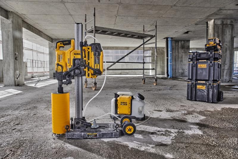 Une machine de forage de carottes DEWALT avec un accessoire de foret cylindrique jaune et un réservoir d'eau connecté est placée sur le sol d'un chantier en béton. À l'arrière-plan, on aperçoit un échafaudage et des boîtes de rangement empilables de marque DEWALT.