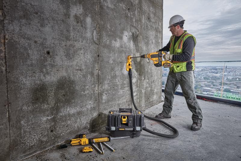 Un ouvrier du bâtiment portant un gilet de sécurité et un casque utilise une perceuse sans fil DEWALT avec un accessoire d’extraction de poussière pour percer un mur en béton. L’ouvrier se tient sur un grand bâtiment avec une vue sur la ville à l’arrière-plan. Plusieurs outils DEWALT et une mallette à outils sont posés au sol à proximité.