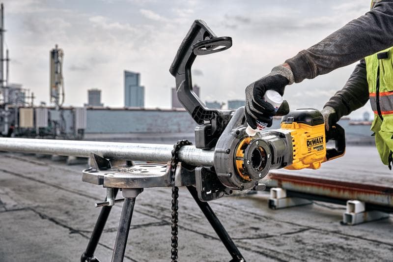 Une personne utilise un outil électrique jaune DEWALT DCE700X2 pour fileter un tuyau en métal fixé sur un support de travail, à l'extérieur sur un toit avec des immeubles visibles en arrière-plan. L'utilisateur porte des gants et un gilet de sécurité.