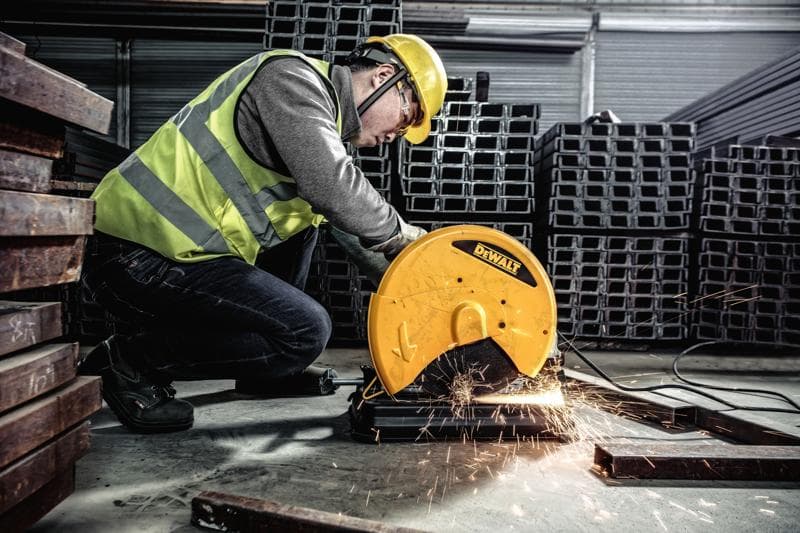 Une personne portant un équipement de sécurité utilise une scie à métaux DEWALT, modèle D28730, pour couper des barres métalliques dans un environnement industriel. Des étincelles jaillissent de la zone de coupe.