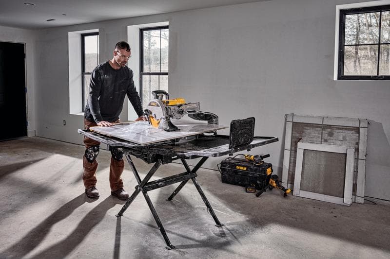 Une personne se tient à côté d’une scie à carrelage DEWALT D36000S installée sur son support dans une pièce en cours de construction. La scie est placée sur un support pliable noir, avec des outils et un sac à outils DEWALT au sol à proximité. La pièce possède des murs non finis, des fenêtres et des matériaux de construction appuyés contre le mur.