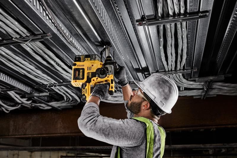 Une personne portant un casque, des lunettes de sécurité, des gants et un gilet de sécurité utilise une perceuse rotative sans fil jaune DEWALT pour percer un plafond métallique avec des câbles électriques.