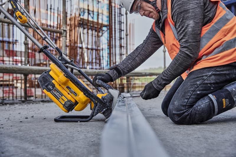 Ouvrier utilise un outil jaune DEWALT pour scier du métal sur chantier, en gilet orange et équipement de sécurité.