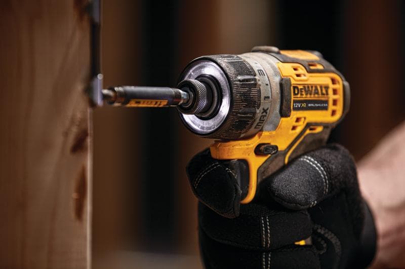Une perceuse sans fil DEWALT visse une vis dans du bois, tenue par une personne avec des gants noirs.
