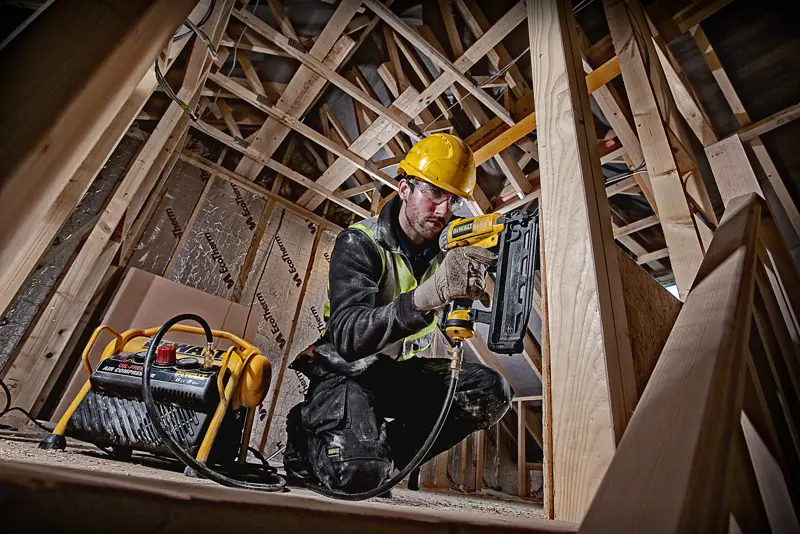 Ouvrier utilise un cloueur DEWALT avec compresseur sur chantier bois, équipé de protections.