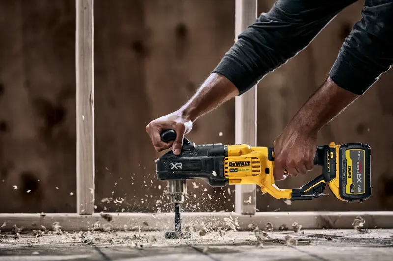 Une personne utilise une perceuse sans fil DEWALT DCD444N avec une poignée latérale pour percer du bois, produisant des copeaux et de la poussière de bois. Une structure de construction est visible en arrière-plan.