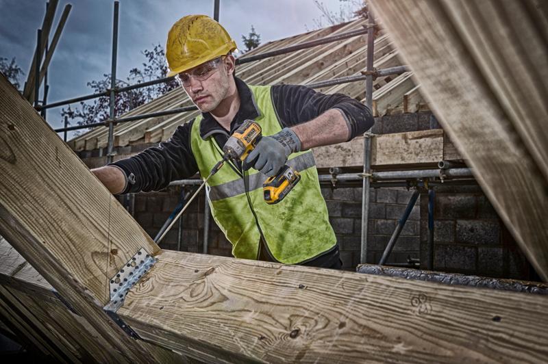 Personne utilisant une perceuse DEWALT sur des poutres en bois, avec un gilet haute visibilité et un casque de sécurité.